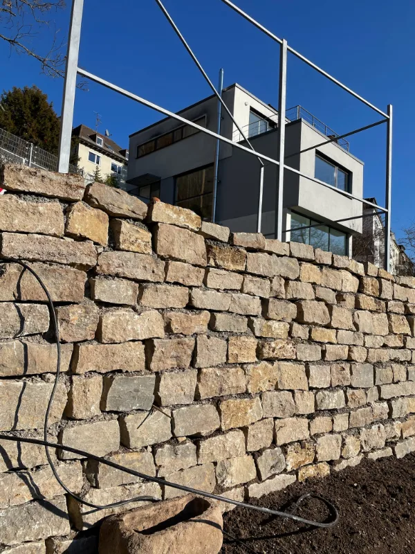 Neu errichtete Stützmauer aus Muschelkalk in lagerhaftem Verband, dahinter das Wohnhaus am Hang